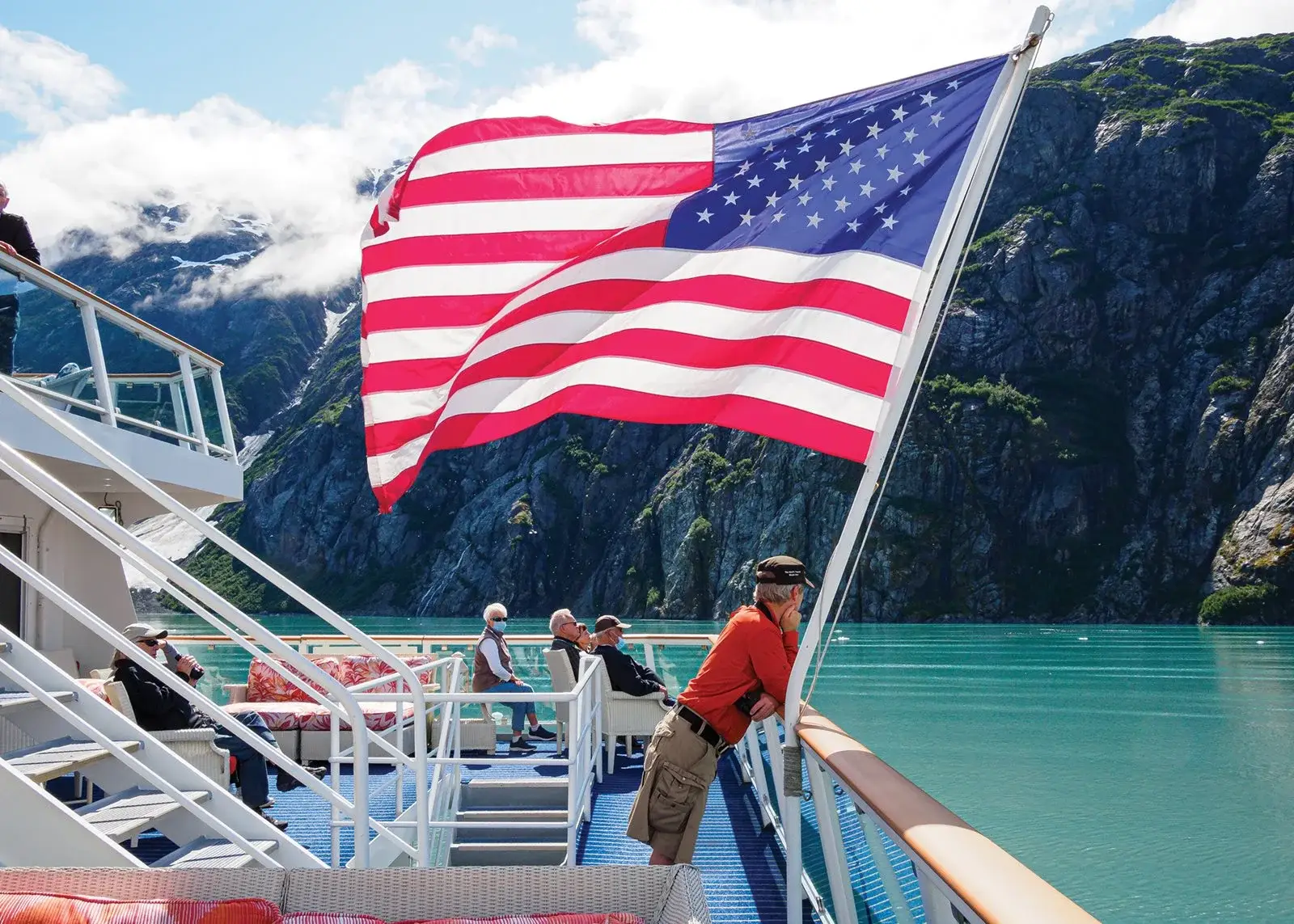 Photo-of-passengers-on-ship-deck-behind-flying-American-flag-near-Alaskan-mountains